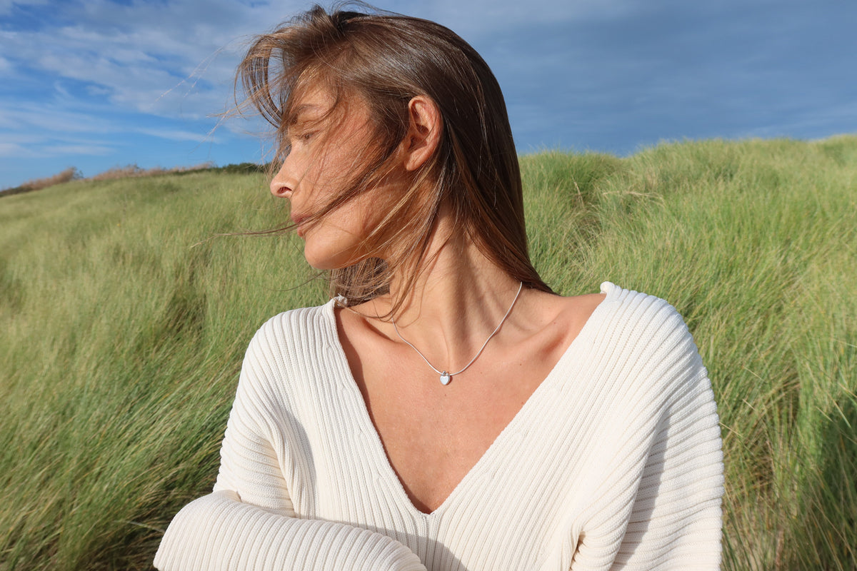 Frau im weißen Strickpullover in den Dünen trägt den silbernen Muttermilch-Anhänger Amore in Herzform.