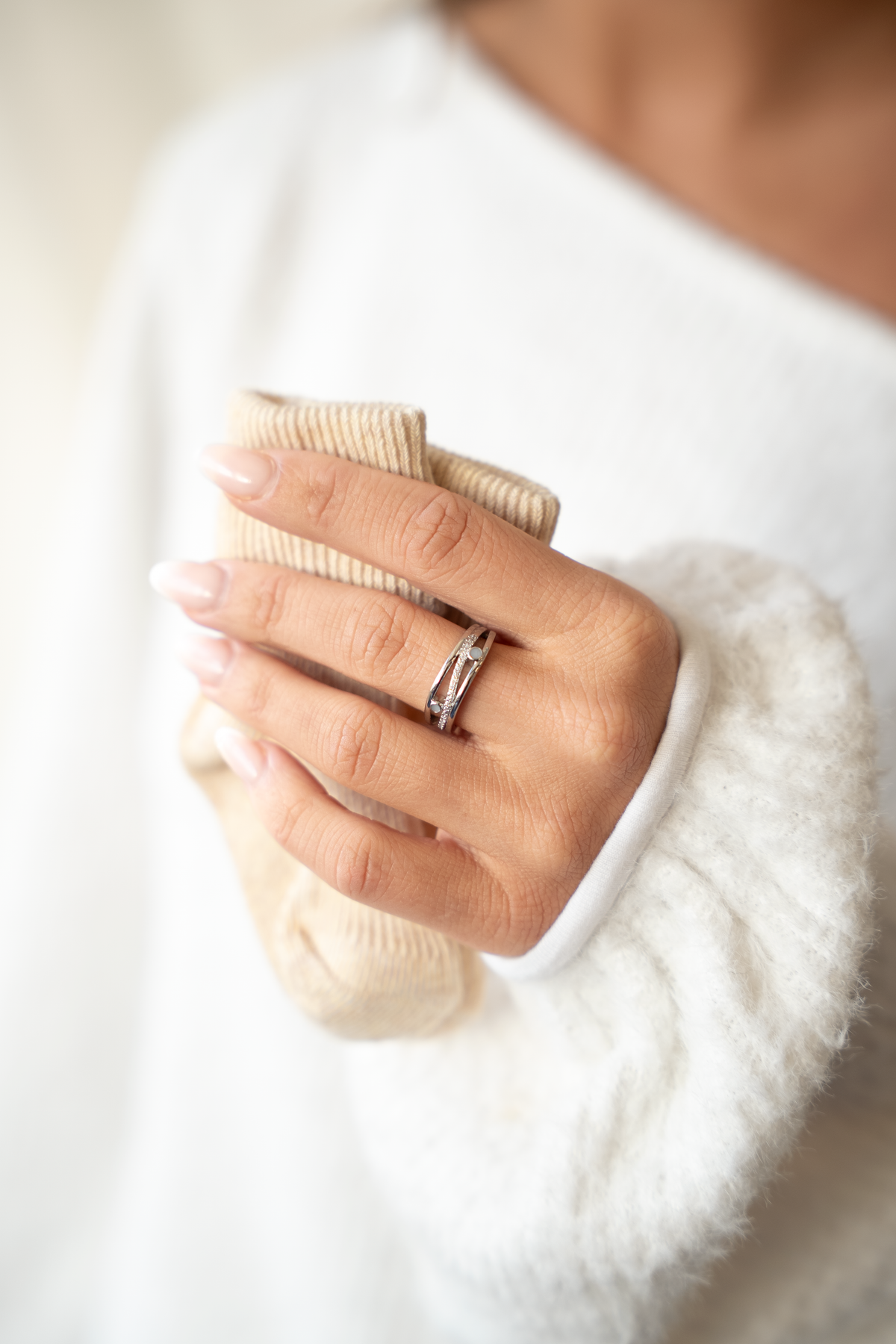 Hand mit beigem Babysöckchen vor weißem Pullover, am Finger der Charlotte Muttermilchring mit drei Bändern