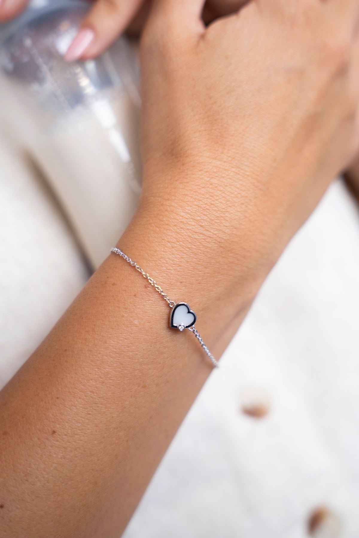 Weißgoldenes Esmée-Armband mit herzförmigem Muttermilchstein am Handgelenk, neben einem Fläschchen Milch.