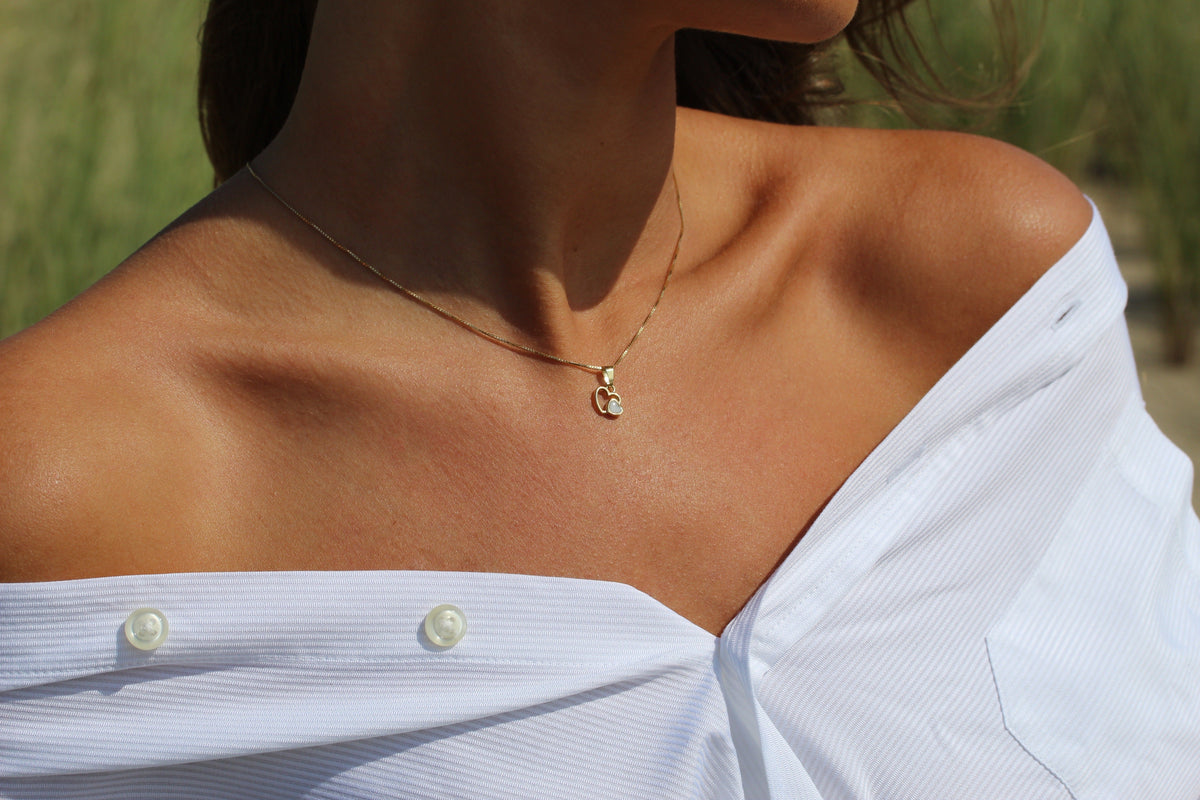 Nahaufnahme von Hals und Schulter mit dem goldenen Heart to Heart Anhänger an feiner Goldkette, weißes Hemd.