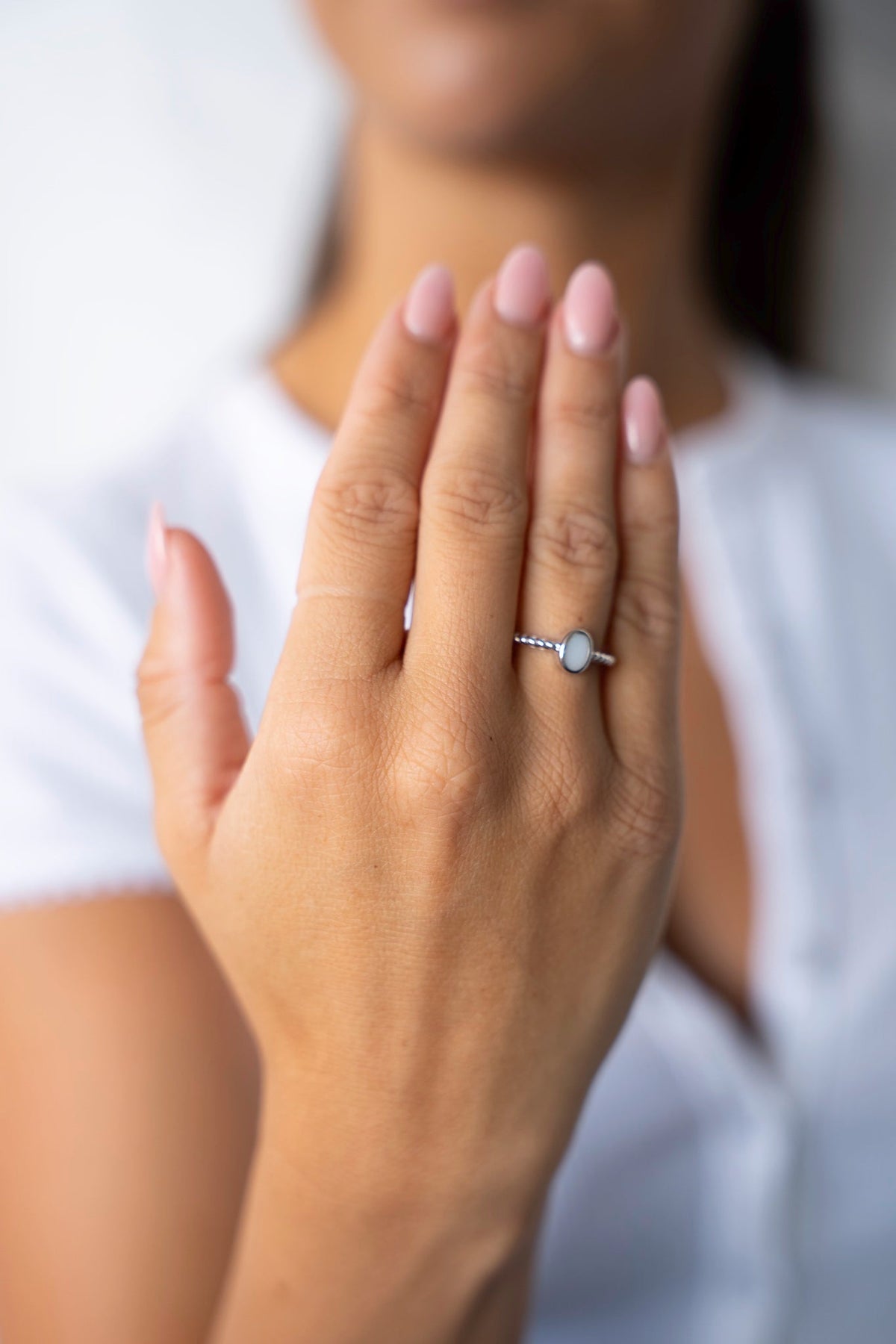 Erhobene Hand mit dem weißgoldenen Ilse-Ring mit ovalem Muttermilchstein, vor einem weißen Shirt.